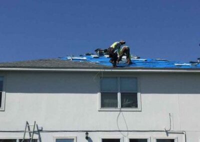A worker in a green shirt is repairing a roof, sitting and using tools on a sunny day. a ladder leans against the two-story house, under a clear blue sky.