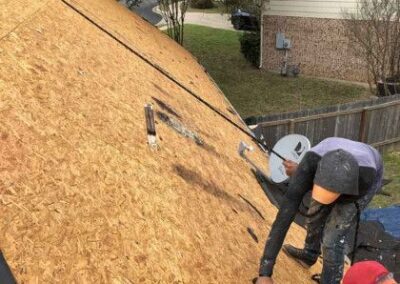 Two workers on a sloping plywood roof: one kneeling with a nail gun, the other standing beside, both repairing or installing the roof, with residential houses and trees in the background.