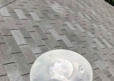 A close-up of a round metal vent on a gray shingled roof, viewed under bright sunlight. the focus is on the textured patterns of the roof and the shiny surface of the vent.