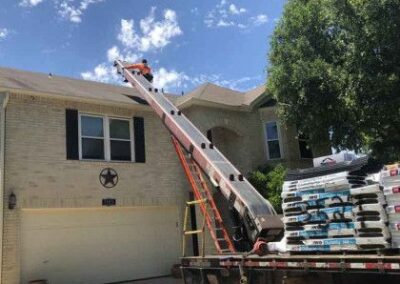 A roofing conveyor belt delivers shingles to the roof of a two-story house, with a flatbed truck loaded with shingle bags parked beside the house under a clear blue sky.