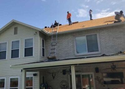 Workers are on the roof of a two-story house, installing shingles under a clear sky. a ladder leans against the house for access.