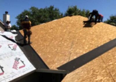 Workers install plywood sheathing on a steep residential roof on a sunny day, with clear blue sky in the background.