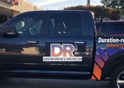 A black truck with a vibrant blue and orange promotional wrap displaying the logo and name of "duration roofing & construction" parked beside a road.