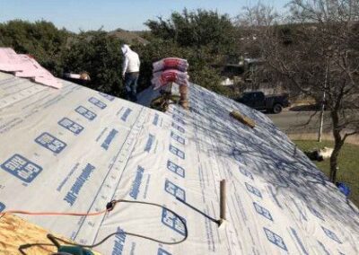 Workers on a steep roof covered with underlayment, installing shingles with tools nearby, in a suburban area on a sunny day.