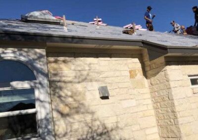 Workers are installing shingles on the roof of a stone house under a clear blue sky, with shadows of tree branches on the house facade.