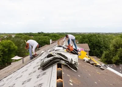 Three workers re-shingling a house roof with tools and materials scattered around, under a cloudy sky, surrounded by a lush green environment.